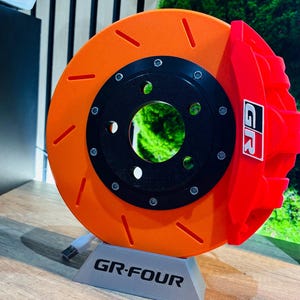 Puede incluir: Un disco de freno naranja y negro con una pinza de freno roja. El disco tiene ranuras y está montado sobre un soporte gris con el texto "GR-FOUR". La pinza tiene las letras "GR".