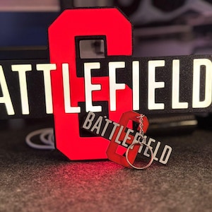 以下が含まれることがあります： 黒い背景に白い文字で「BATTLEFIELD」と書かれた赤い数字の8。同じデザインのキーホルダーも付いています。これらのアイテムは、ビデオゲームに関連している可能性があります。