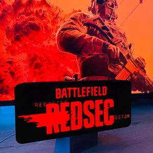 May include: A black sign with the words "Battlefield REDSEC" in red, with a soldier in the background. The background is a fiery orange and red scene with explosions and fighter jets.