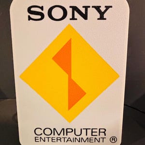 以下が含まれることがあります： 「SONY」という黒い文字が書かれた白い看板、オレンジ色のS字が描かれた黄色のひし形、そして黒い文字で「COMPUTER ENTERTAINMENT®」と書かれている