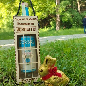 May include: A wooden box with a blue bottle of Zorbas Ouzo inside. The box has a message on the front that says "Let me out! I know you want me!" and the date "30.05.2024". A gold chocolate bunny is sitting in front of the box.