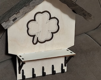 May include: Wooden key holder shaped like a house with a four-leaf clover design and five hooks. The house is painted brown and the clover is white.