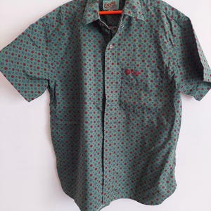 Può includere: Una camicia verde a maniche corte con bottoni e un motivo floreale rosso e blu. La camicia ha una tasca sul petto sinistro con il testo "Crocker" ricamato.
