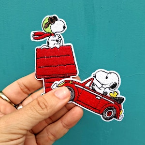 May include: Embroidered patches featuring Snoopy in a red doghouse and driving a red convertible. Snoopy is wearing a green aviator helmet and red scarf. The patches are white, red, black, and yellow.