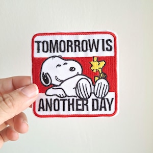Op de afbeelding: Een vierkante patch met een rode rand en de tekst "TOMORROW IS ANOTHER DAY". De patch toont het stripfiguur Snoopy in het wit, liggend met een gele bloem. De patch wordt door een hand vastgehouden.