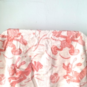 Pode incluir: Uma manta de lã rosa e branca com um padrão de animais de desenho animado rosa correndo e brincando com uma bola. A manta tem uma borda rosa.