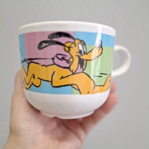 Puede incluir: Una taza de cerámica blanca con una imagen de dibujos animados de un perro corriendo. El perro es amarillo con orejas negras y un collar rosa. El fondo es un mosaico de rosa, azul, verde y amarillo.