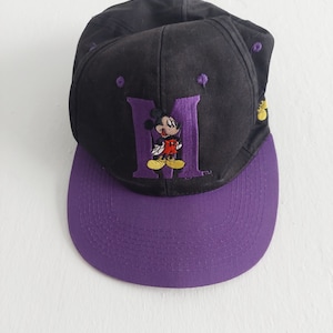 Peut inclure: Casquette de baseball noire et violette avec une lettre "M" violette et une image de dessin animé de Mickey Mouse au centre.