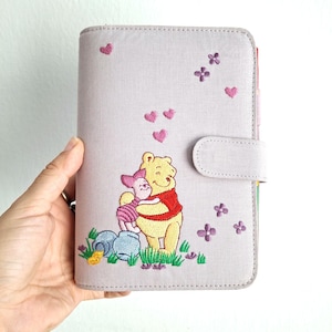 Może przedstawiać: Jasnofioletowy planer z haftowanym wizerunkiem Kubusia Puchatka i Prosiaczka obejmujących się. Planer posiada zapięcie na zakładkę i jest ozdobiony haftowanymi sercami i kwiatami.