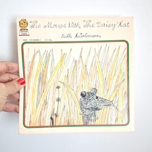 Pode incluir: Um livro infantil vintage intitulado "The Mouse With The Daisy Hat" de Ruth Hürlimann. A capa apresenta uma ilustração de um rato na erva alta. O livro tem uma borda verde e vermelha e o logotipo Picture Lions.