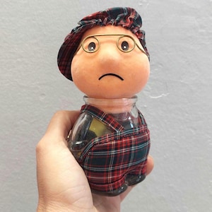 Könnte beinhalten: Eine Puppe aus rotem und grünem kariertem Stoff mit einem Glaskörper, die eine Brille und einen karierten Hut trägt. Die Puppe hat einen traurigen Gesichtsausdruck.