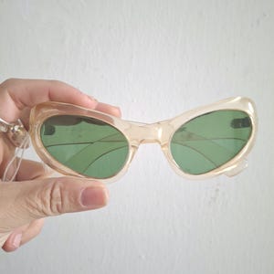 Może przedstawiać: Okulary przeciwsłoneczne w stylu vintage z kocimi oczami, z jasnobrązową oprawką i zielonymi soczewkami. Okulary w stylu retro mają unikalny kształt i klasyczny design. Te okulary przeciwsłoneczne to stylowy dodatek do każdego stroju.