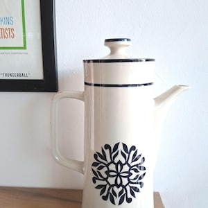 May include: A tall, off-white ceramic coffee pot with a lid and handle. The pot features a dark blue floral design and dark blue stripes around the top. The pot is sitting on a wooden surface.