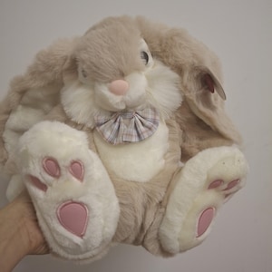 Pode incluir: Um coelho de pelúcia com orelhas longas e caídas e uma gravata borboleta xadrez. O coelho tem pelo bege macio, detalhes brancos e detalhes rosa no nariz e nas patas. O bicho de pelúcia foi projetado para ser fofo.
