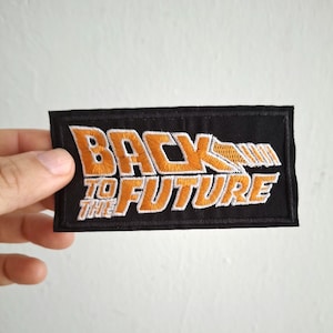 Peut inclure: Un écusson brodé noir avec le texte "Back to the Future" en lettres orange et blanches. L'écusson a une flèche blanche pointant vers la droite.
