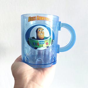 Peut inclure: Une tasse en plastique bleue avec une image de Buzz l'Éclair de Toy Story. La tasse a une poignée et dit "Buzz Lightyear" sur le devant.