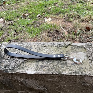 May include: A black leather dog leash with a silver clip. The leash is lying on a gray concrete surface.