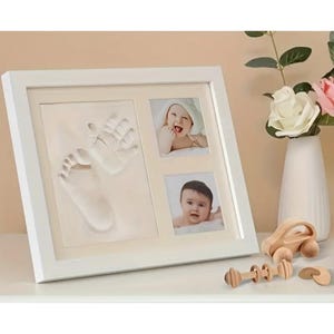Könnte beinhalten: Ein weiß gerahmtes Baby-Andenken mit Hand- und Fußabdruck sowie zwei Babyfotos. Ein Spielzeugauto aus Holz, eine Holzrassel und eine Vase mit Blumen stehen auf dem Tisch.