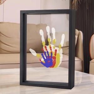 Könnte beinhalten: Ein gerahmtes Kunstwerk mit bunten Handabdrücken. Die Handabdrücke sind in Gelb, Rot, Blau und Weiß gehalten und überlagert, um ein familienbezogenes Design zu schaffen. Der Rahmen ist schwarz und rechteckig, mit einer klaren Glasfront.
