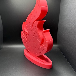Puede incluir: Un rompecabezas tridimensional de color rojo brillante con forma de llama, que se mantiene en posici&oacute;n vertical sobre una base ovalada. El rompecabezas est&aacute; compuesto por m&uacute;ltiples piezas entrelazadas que forman la silueta de un fuego.
