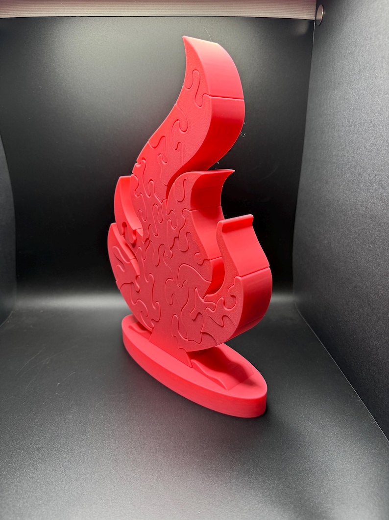 Puede incluir: Un rompecabezas tridimensional de color rojo brillante con forma de llama, que se mantiene en posici&oacute;n vertical sobre una base ovalada. El rompecabezas est&aacute; compuesto por m&uacute;ltiples piezas entrelazadas que forman la silueta de un fuego.