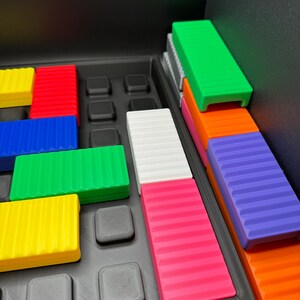May include: A colorful puzzle game with rectangular blocks in various colors, including green, red, blue, yellow, white, pink, orange, and purple. The blocks are arranged in a black tray with square indentations.