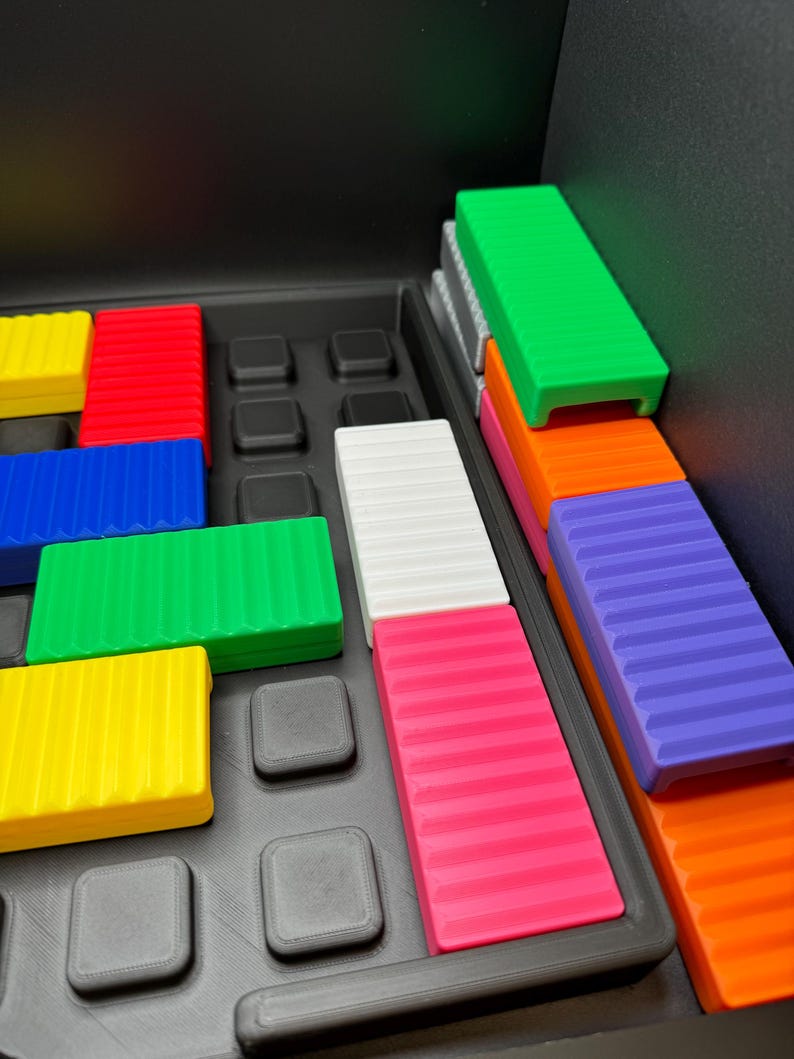 May include: A colorful puzzle game with rectangular blocks in various colors, including green, red, blue, yellow, white, pink, orange, and purple. The blocks are arranged in a black tray with square indentations.