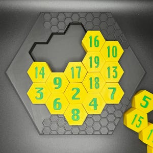 Puede incluir: Un rompecabezas hexagonal amarillo con n&uacute;meros verdes del 1 al 19. Al rompecabezas le falta una pieza. La pieza que falta se muestra a la derecha del rompecabezas.