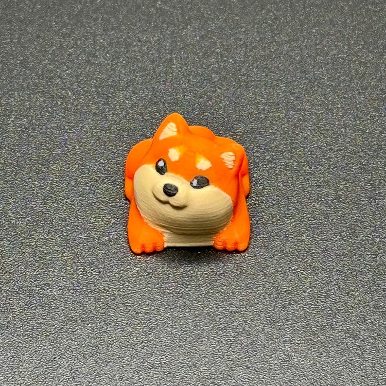 May include: A small, orange and white Shiba Inu dog figurine. The dog has black eyes, a black nose, and a smiling expression. The figurine is resting on a dark gray surface.
