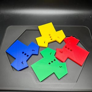 Puede incluir: Cuatro piezas de juego geom&eacute;tricas de colores azul, amarillo, rojo y verde. Cada pieza tiene una forma &uacute;nica con tres agujeros circulares y una peque&ntilde;a extensi&oacute;n rectangular. Est&aacute;n dispuestas sobre una superficie gris oscuro.