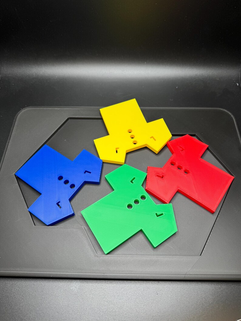 Puede incluir: Cuatro piezas de juego geom&eacute;tricas de colores azul, amarillo, rojo y verde. Cada pieza tiene una forma &uacute;nica con tres agujeros circulares y una peque&ntilde;a extensi&oacute;n rectangular. Est&aacute;n dispuestas sobre una superficie gris oscuro.