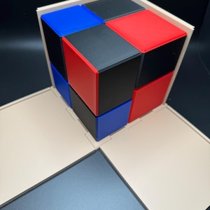 May include: A 3x3 cube made of smaller cubes, each with a different color: red, blue, and black. The cube is sitting inside a beige box with the lid open.
