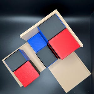 May include: A set of five wooden blocks in various colors, including black, red, and blue. The blocks are arranged in a geometric pattern on a beige surface.