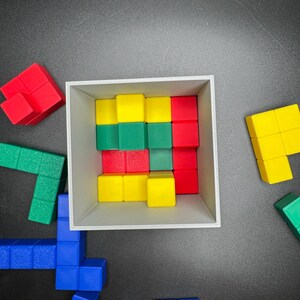 Puede incluir: Una caja blanca llena de bloques de madera de colores. Los bloques est&aacute;n dispuestos en un patr&oacute;n y son rojos, verdes y amarillos. La caja est&aacute; sobre una superficie negra.