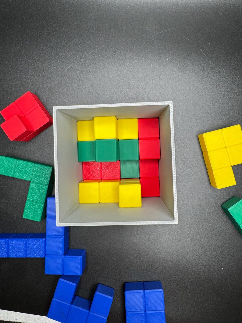 Puede incluir: Una caja blanca llena de bloques de madera de colores. Los bloques est&aacute;n dispuestos en un patr&oacute;n y son rojos, verdes y amarillos. La caja est&aacute; sobre una superficie negra.