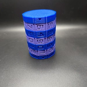 May include: A cylindrical puzzle toy in shades of blue and purple. The toy is composed of stacked, segmented rings with numbers printed on the sides. The top is a solid blue cylinder. The puzzle sits on a dark surface.