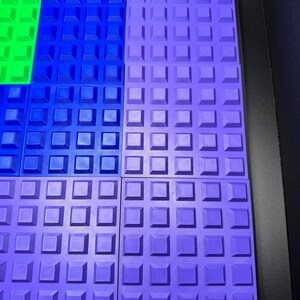 May include: A close-up of several colorful, textured, square building blocks. The blocks are arranged in a grid pattern, with sections of green, blue, and purple. Each block has a raised square pattern.