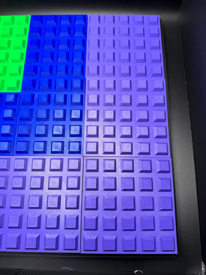 May include: A close-up of several colorful, textured, square building blocks. The blocks are arranged in a grid pattern, with sections of green, blue, and purple. Each block has a raised square pattern.