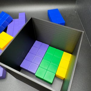 May include: A black rectangular container holds colorful, geometric puzzle pieces. The pieces are various colors, including blue, purple, green, and yellow. The pieces are arranged inside the container and scattered around it.