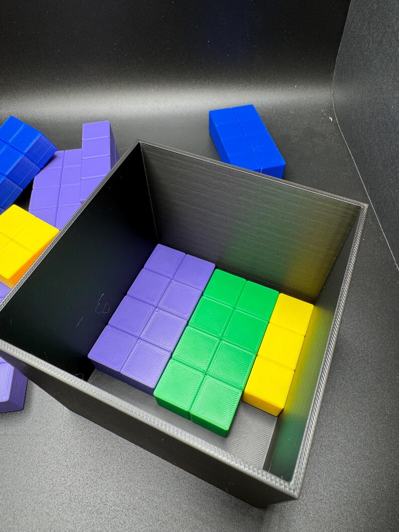 May include: A black rectangular container holds colorful, geometric puzzle pieces. The pieces are various colors, including blue, purple, green, and yellow. The pieces are arranged inside the container and scattered around it.