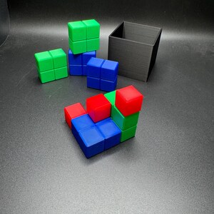 May include: A collection of colorful cube puzzles in red, green, and blue, arranged in various configurations. A black square container sits in the background. The cubes are made of plastic and are on a dark gray surface.