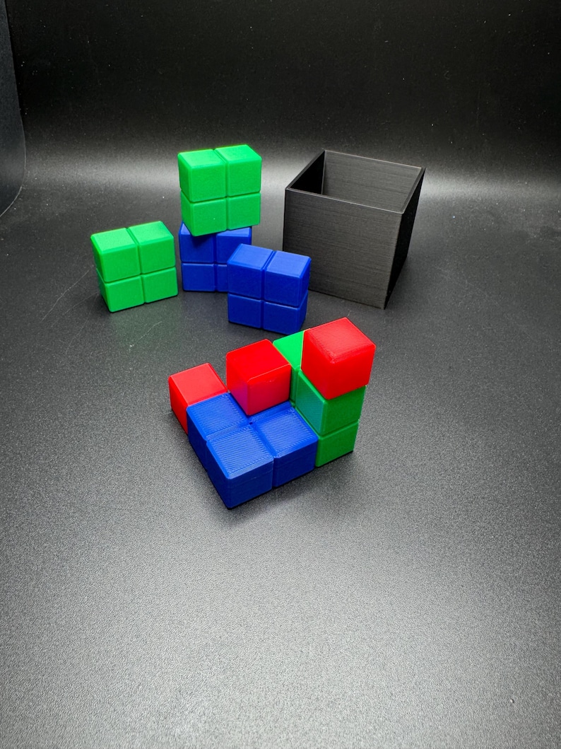 May include: A collection of colorful cube puzzles in red, green, and blue, arranged in various configurations. A black square container sits in the background. The cubes are made of plastic and are on a dark gray surface.