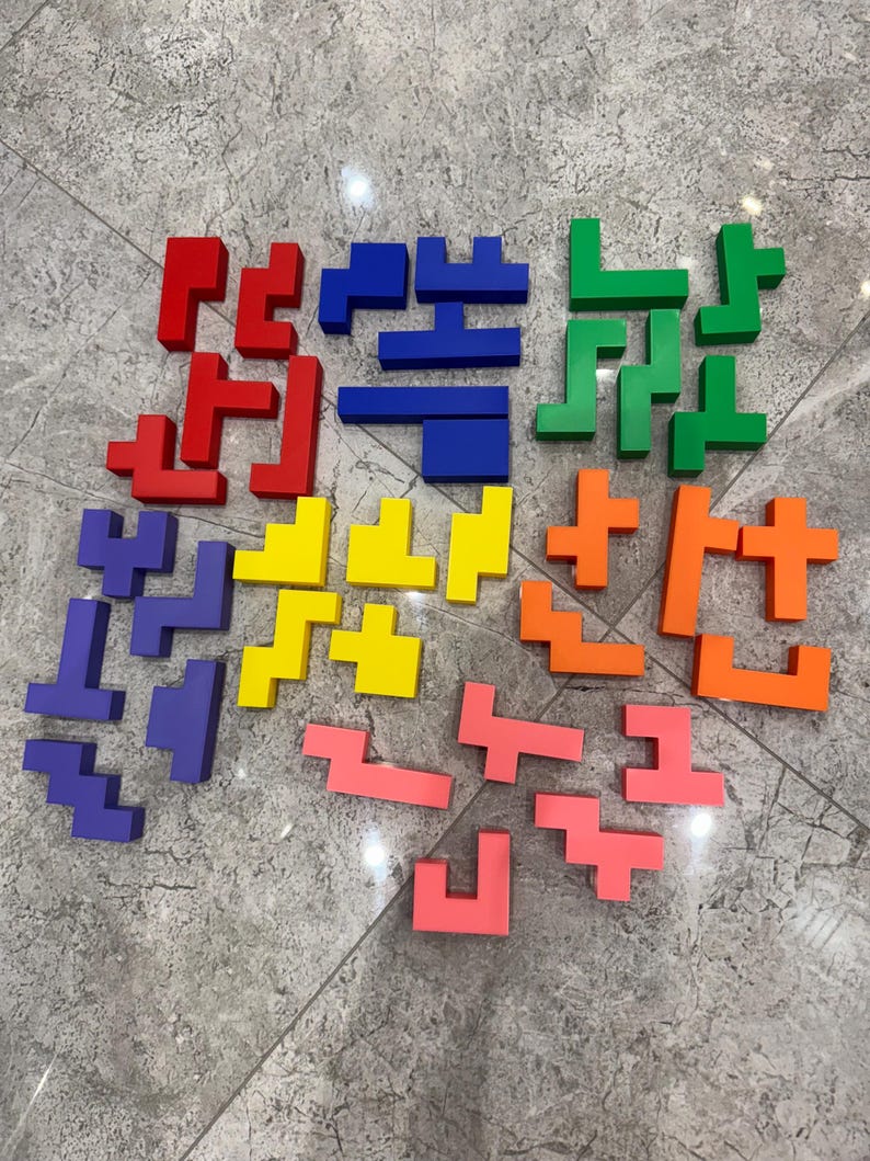 May include: A set of colourful Tetris-like puzzle pieces in red, blue, green, yellow, and orange. The pieces are made of plastic and are arranged in a circular pattern on a grey tiled floor.