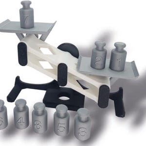 May include: A white and black seesaw toy with gray plastic weights numbered 1 through 7. The seesaw is balanced with weights 2 and 1 on one side and weight 7 on the other side. There are six additional weights on the ground, numbered 6, 4, 3, 5, 5, and 3.