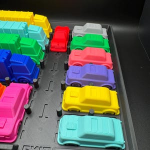 May include: A selection of brightly coloured toy cars, including red, green, and blue, displayed on a black tray. The cars are all the same size and shape, with a simple, blocky design. The word "EXIT" is visible on the tray.