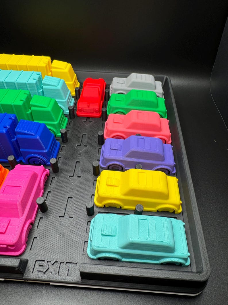 May include: A selection of brightly coloured toy cars, including red, green, and blue, displayed on a black tray. The cars are all the same size and shape, with a simple, blocky design. The word "EXIT" is visible on the tray.