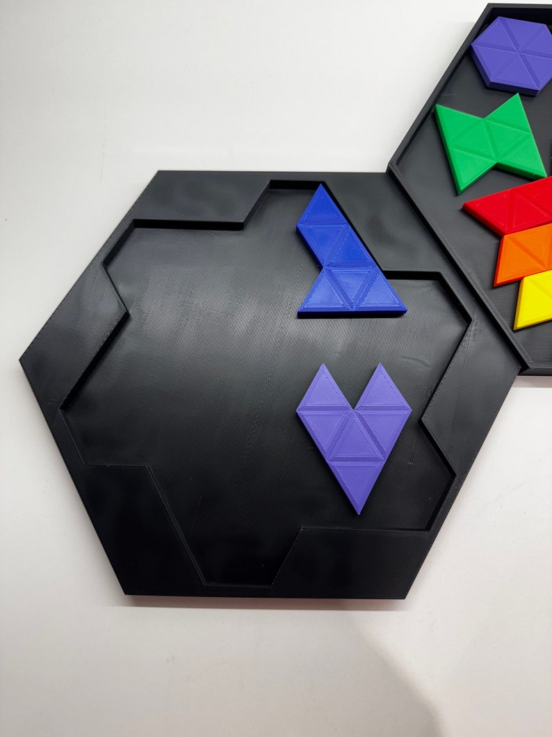Op de afbeelding: Een zwart zeshoekig puzzelbord met een paars hartvormig stuk en een blauw driehoekig stuk op zijn plaats. Het bord heeft een verhoogde rand en is gemaakt van plastic.