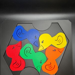 May include: A colorful wooden puzzle featuring six duck-shaped pieces in red, blue, yellow, green, and orange. The pieces fit into a black hexagonal frame, with each duck having a distinct design.