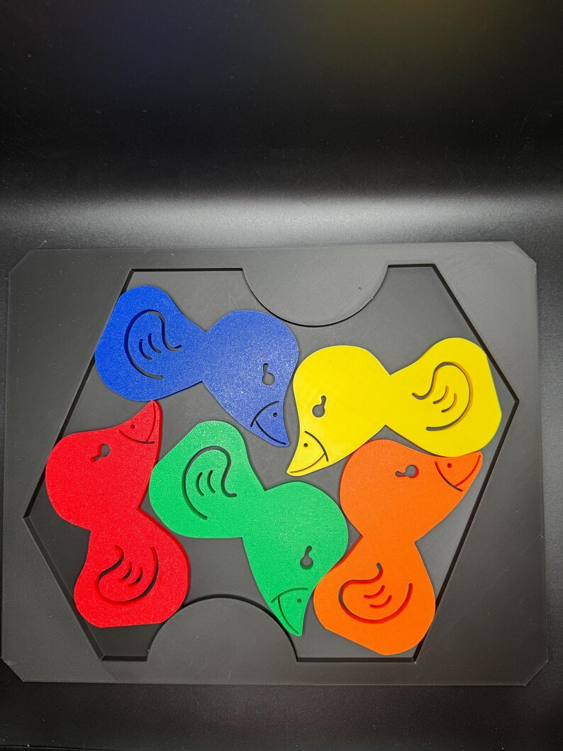 May include: A colorful wooden puzzle featuring six duck-shaped pieces in red, blue, yellow, green, and orange. The pieces fit into a black hexagonal frame, with each duck having a distinct design.
