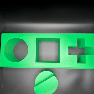 Op de afbeelding: Een felgroen rechthoekig dienblad met drie verschillende uitsparingen: een cirkel, een vierkant en een plusteken. Onder het dienblad ligt een apart felgroen rond onderdeel met een verhoogde rechthoekige balk over het midden.